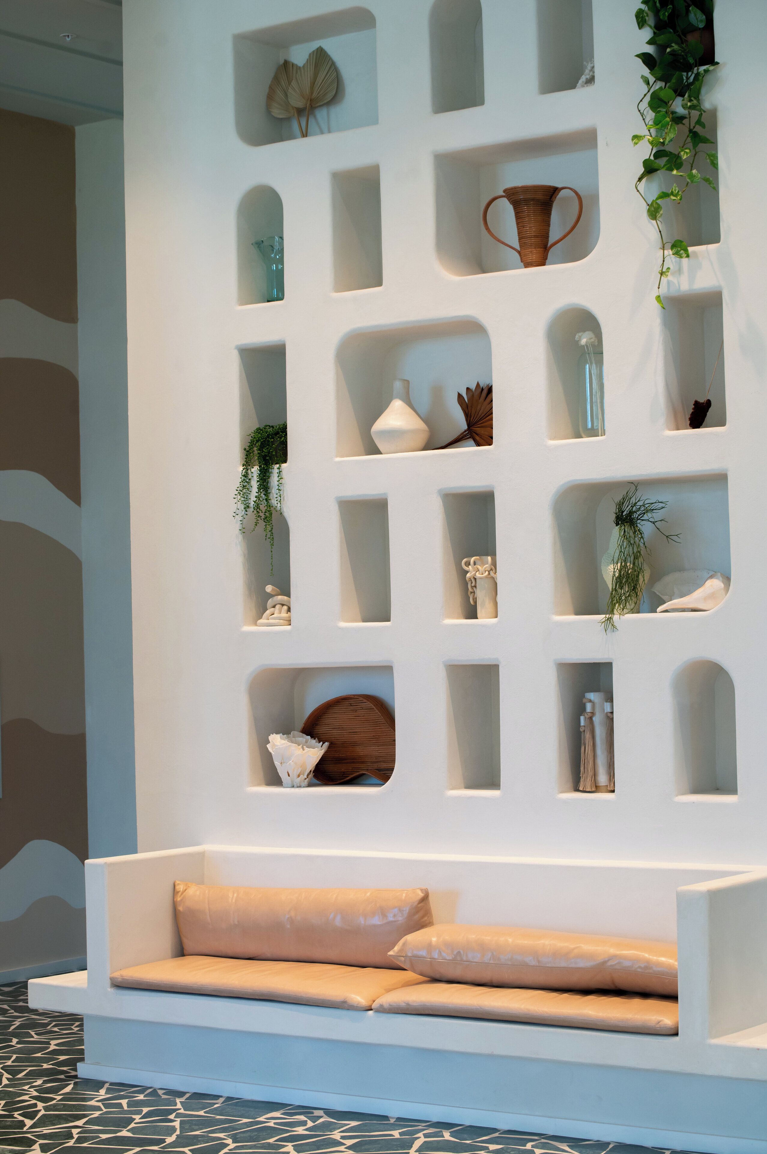 A modern interior with a white wall featuring a grid of shelves displaying various decorative items and a white bench with cushions.