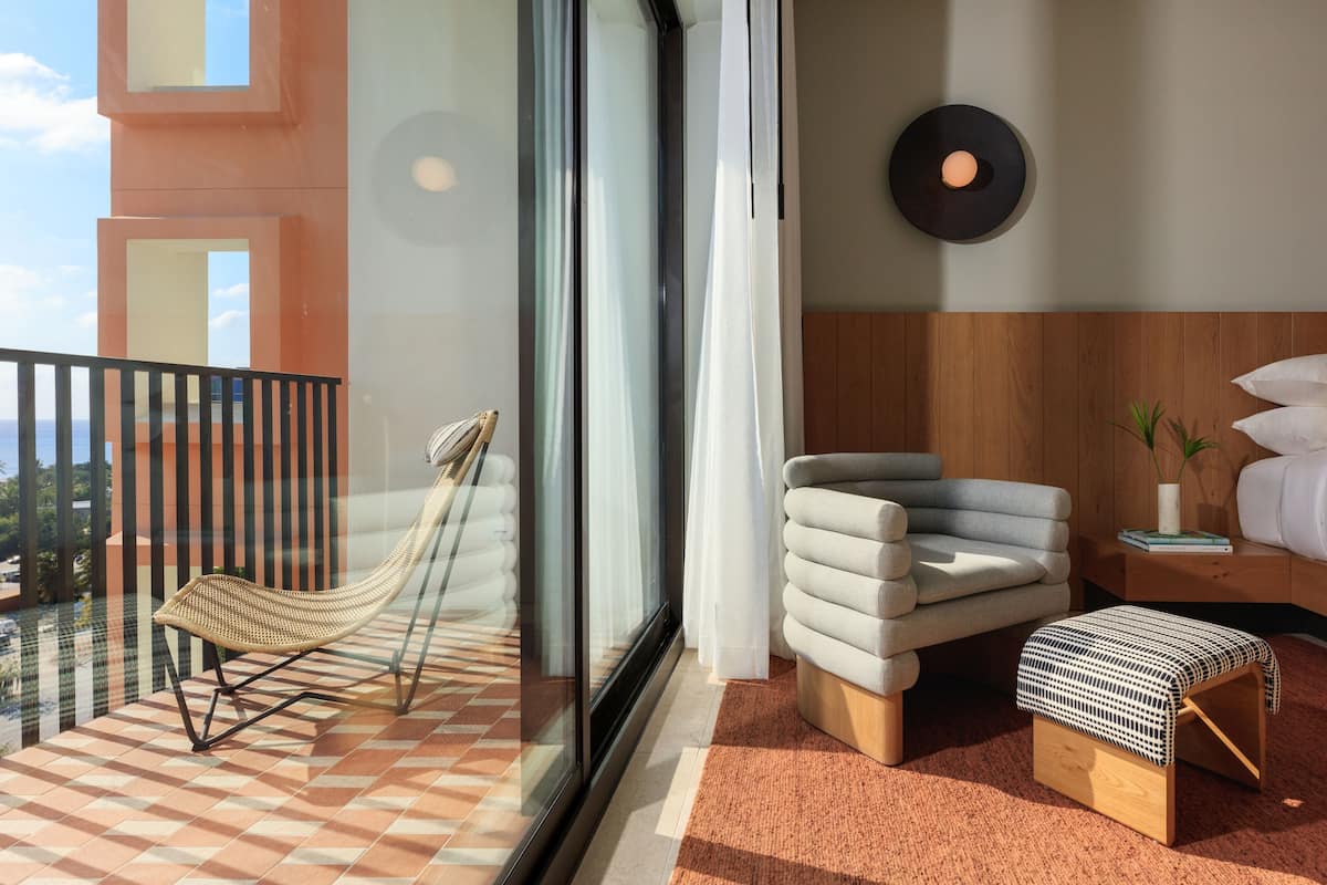 A balcony with a hammock, a cushioned chair, a bedside table with a lamp, and a bed with pillows.