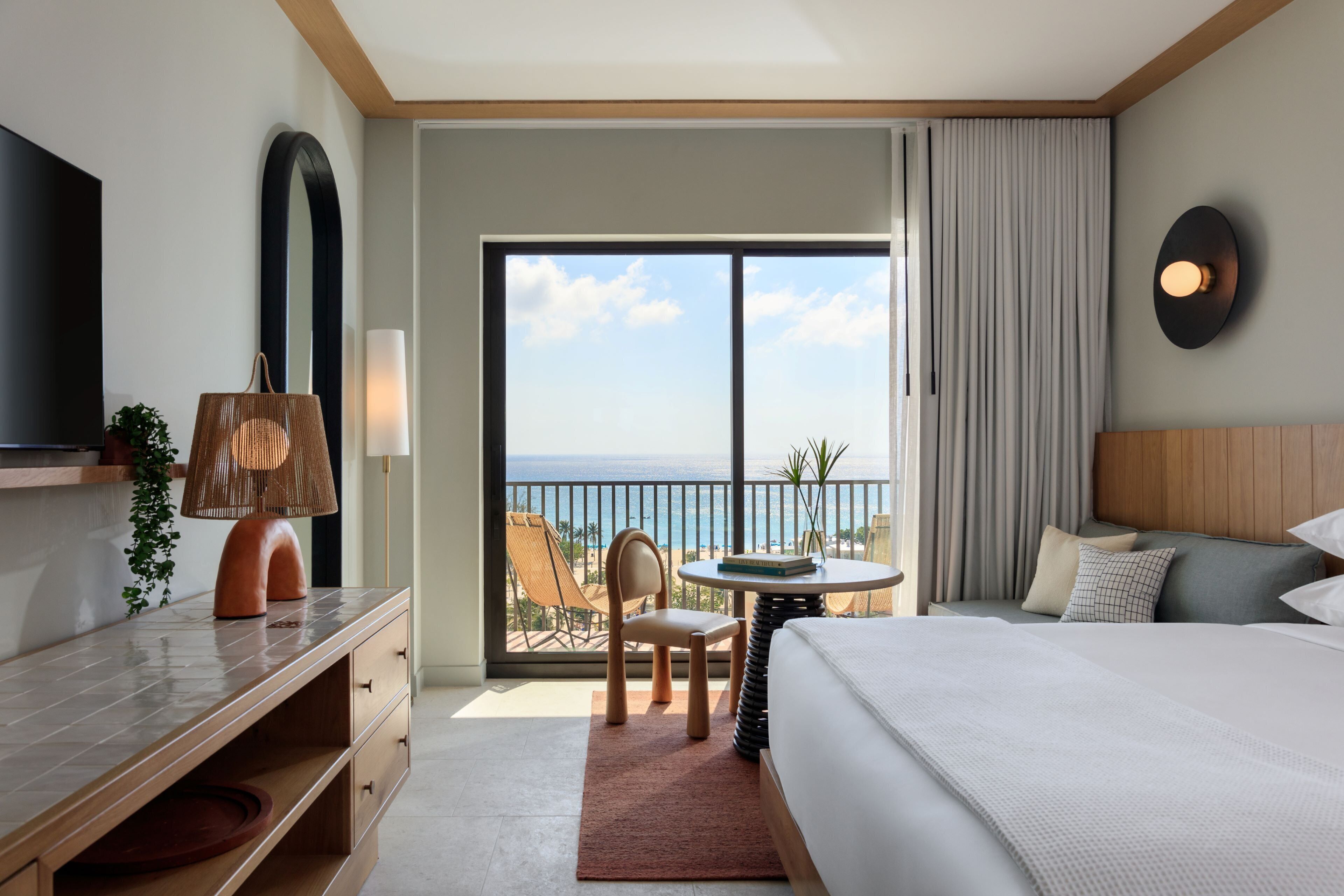 Une chambre d’hôtel comprenant un lit, un bureau, une chaise, une table, un téléviseur, une grande fenêtre donnant sur l’océan et un balcon.