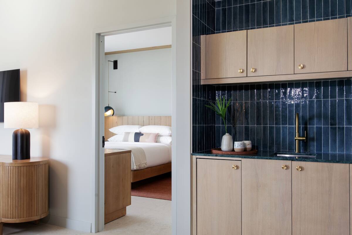 A modern hotel room with a wooden bed, a bedside table with a lamp, a bathroom with a blue-tiled wall, and wooden cabinets.