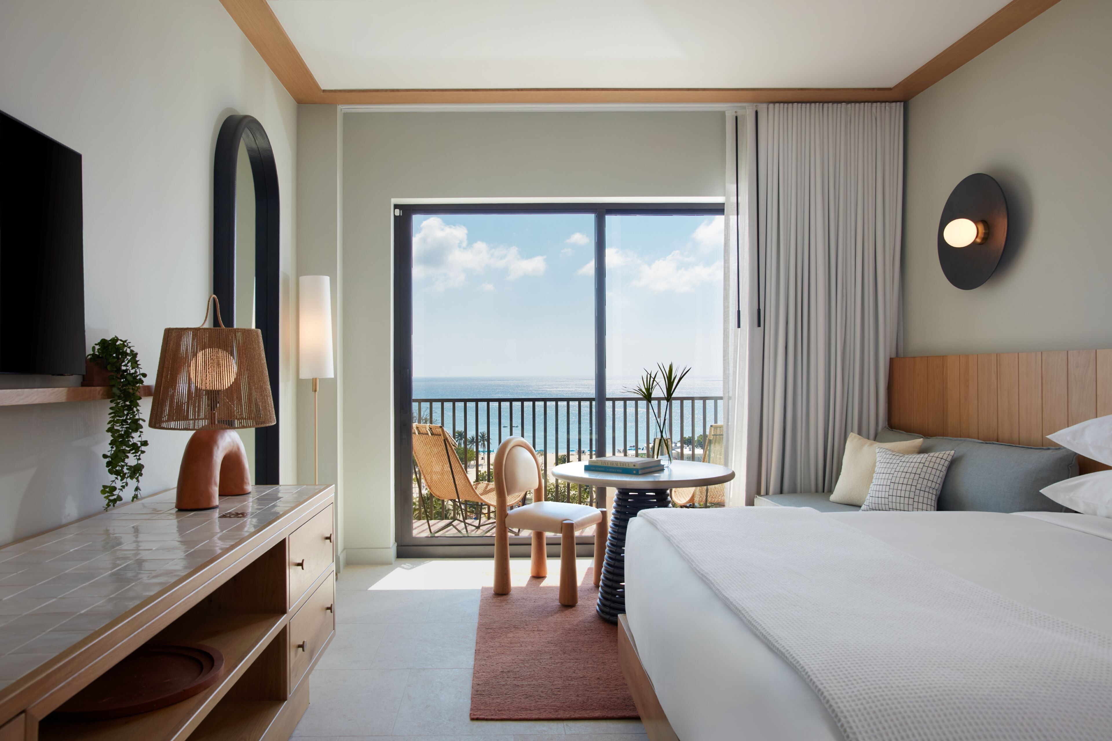 A hotel room with a bed, a desk, a chair, a table, a TV, and a balcony with a view of the ocean.