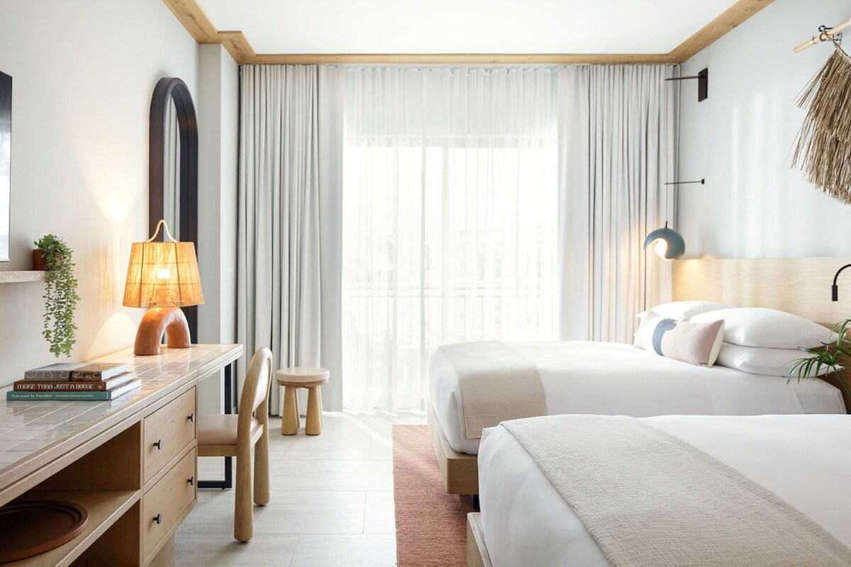 Habitación de hotel con escritorio, dos camas, una silla, un espejo grande y ventana con cortinas.