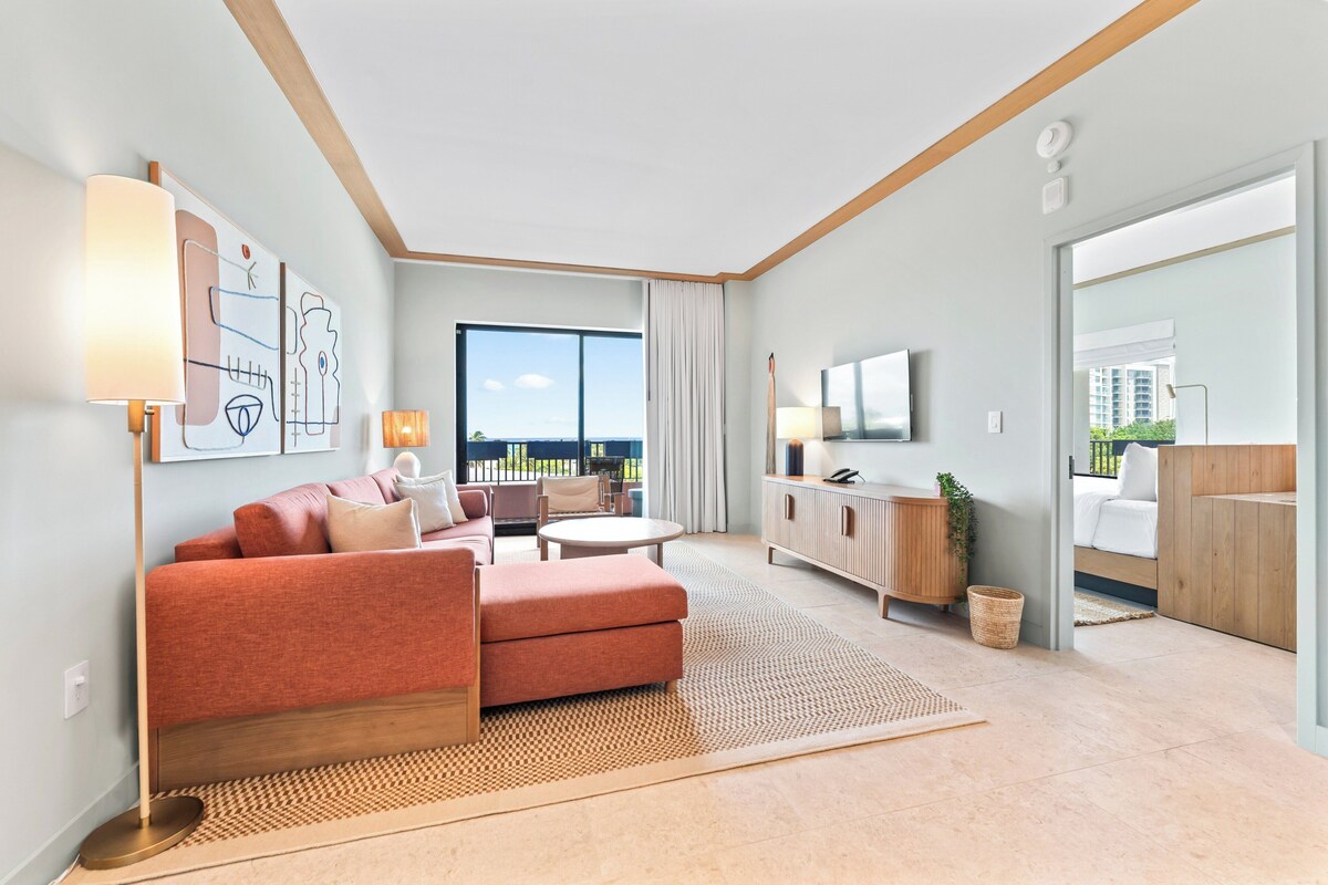 Una habitación de hotel moderna con sofá, mesa de centro, televisor, ventana grande y balcón.