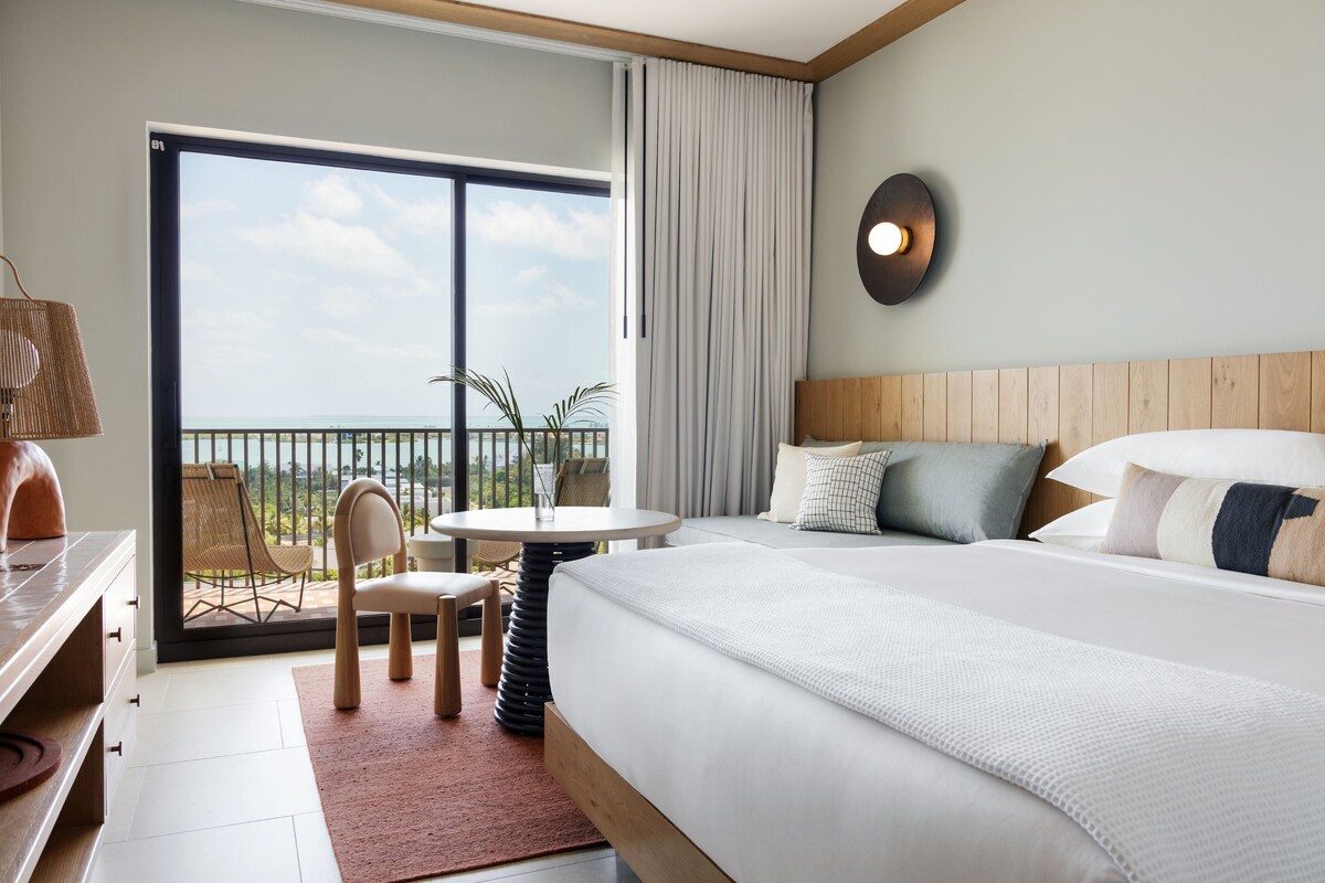 Habitación de hotel con una cama grande, una mesa redonda, dos sillas, vista al mar y un balcón con barandilla.