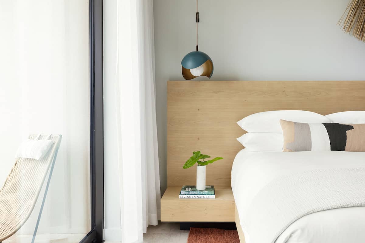 A modern bedroom with a wooden headboard, a bedside table with a plant and books, a hanging pendant light, and a wicker chair.