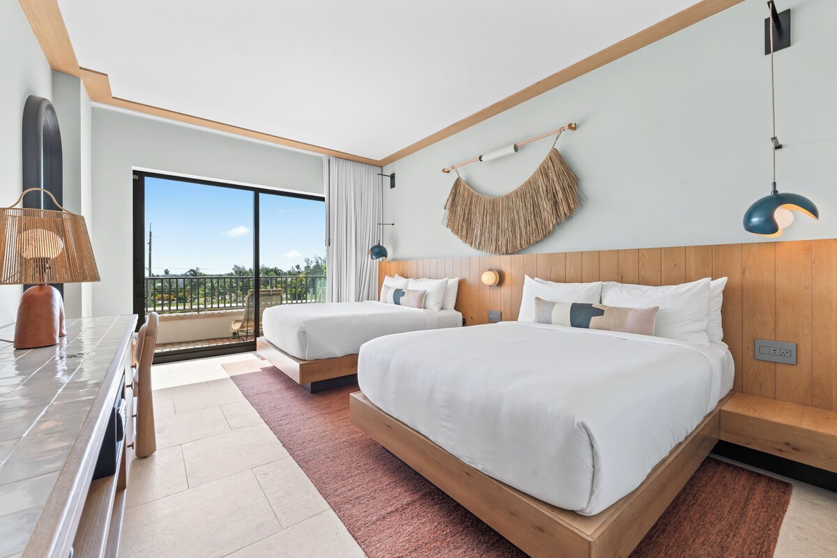Una habitación de hotel moderna con dos camas, un ventanal grande, cabecero de madera y vistas al exterior.