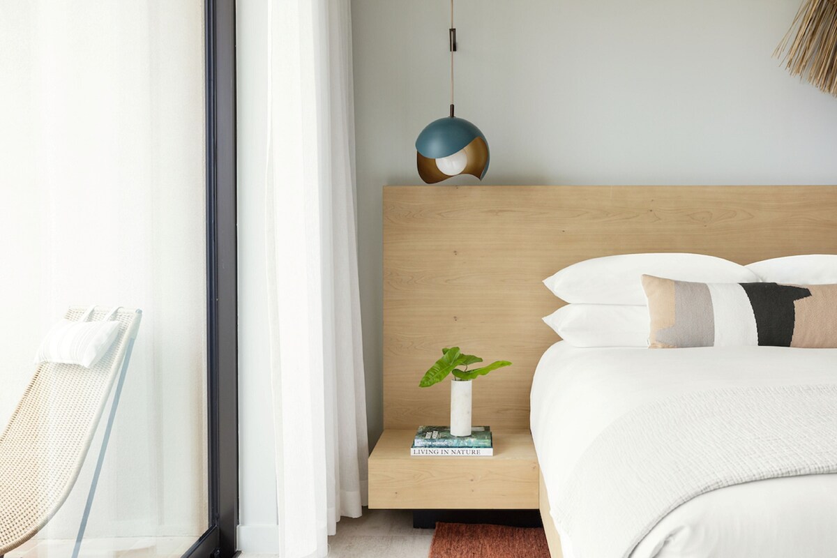 Un dormitorio moderno con cabecera de madera, una mesita de noche con una planta y libros, una lámpara colgante y un amplio ventanal con cortinas.