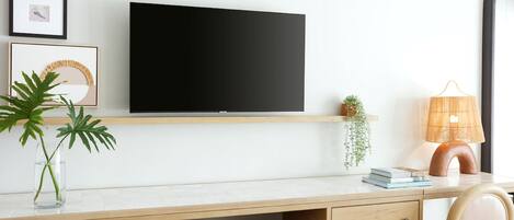 A modern room with a flat-screen TV mounted on the wall, a wooden desk with a chair, a small plant, and a lamp.