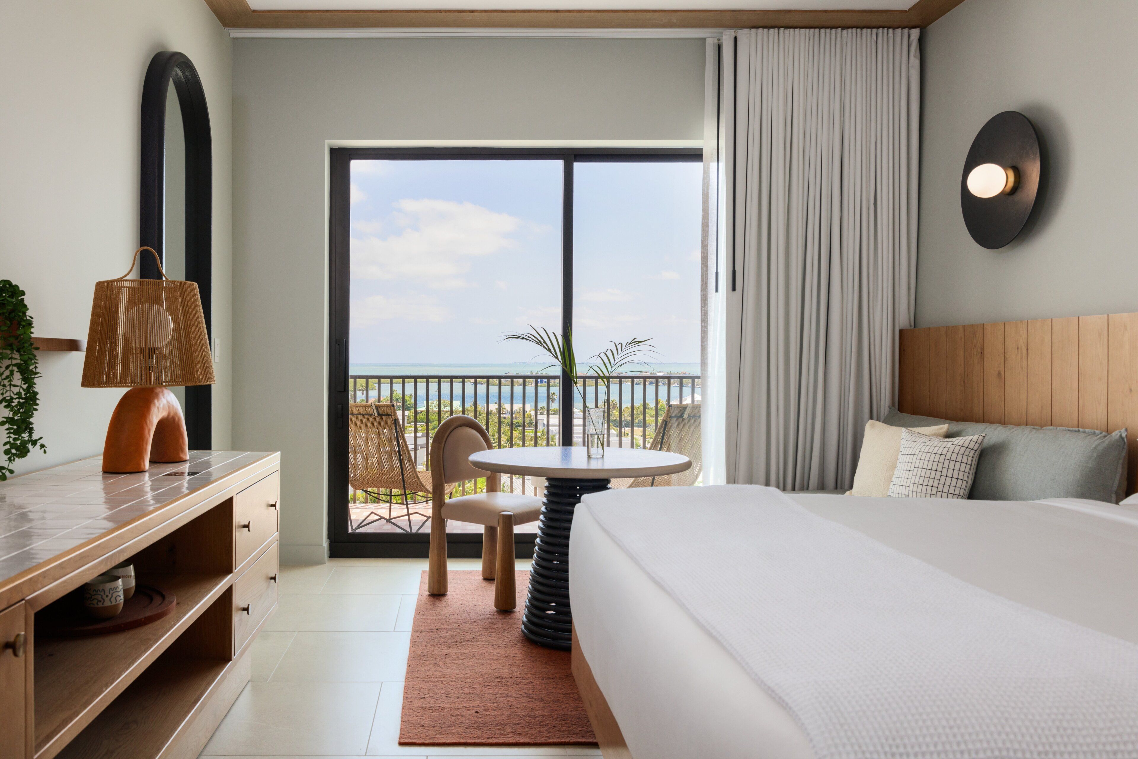 Habitación de hotel con cama, escritorio, silla, mesa y vista al mar a través de un ventanal grande.