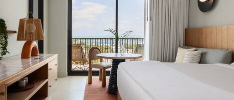 Habitación de hotel con cama, escritorio, silla, mesa y vista al mar a través de un ventanal grande.