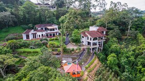 A large, white, two-story house with a red roof, surrounded by lush greenery and a small swimming pool.