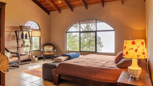 A bedroom with a large arched window, a bed with a brown bedspread, a wooden ceiling, and a lamp on a bedside table.