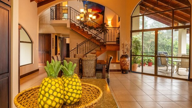 A spacious living area with a staircase, a dining table with chairs, and a large window overlooking greenery.