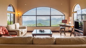 A spacious living room with a large arched window offering a mountain view, a comfortable sofa, a coffee table, and a TV.