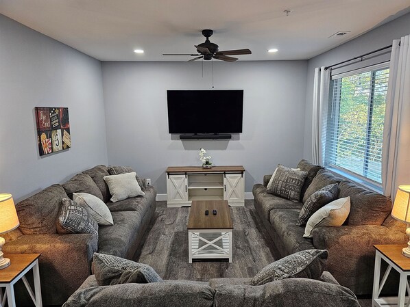 A living room with two sofas, a coffee table, a TV mounted on the wall, and a ceiling fan.