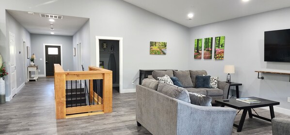 A modern living room with a grey sofa, a wooden staircase, and a flat-screen TV mounted on the wall. - Heart of Branson Retreat / Hot Tub / Pool Table / Game Room / Fire Pit / Lvl2 EV (Branson)