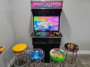 Une borne d’arcade proposant une sélection de jeux classiques, deux tabourets aux motifs colorés et un panneau fixé au mur.