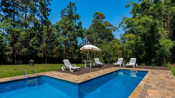 Una piscina rectangular con borde de piedra, rodeada de sillas reclinables blancas y una mesa con sombrilla.