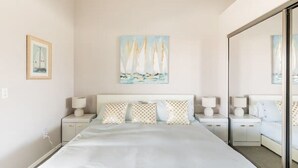 A bedroom with a large bed, two bedside tables with lamps, a painting of sailboats, and a mirror on the wall.