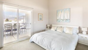 A bedroom with a large bed, a painting of sailboats, a dining area with chairs and a table, and a sliding glass door leading to an outdoor patio.