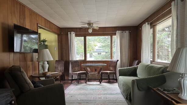 Una sala de estar acogedora con paredes de madera, un ventilador de techo, un televisor montado en la pared, una mesa de comedor con sillas y un sofá.