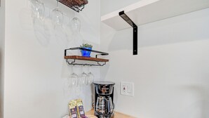 Una cocina con cafetera, un estante con cristalería y una bandeja de cápsulas de café.