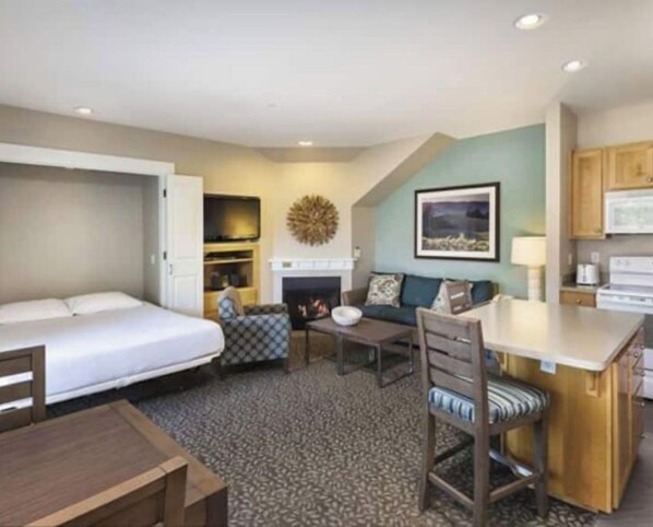 A compact hotel room with a bed, a sofa, a small table, a chair, a fireplace, and a kitchen area.