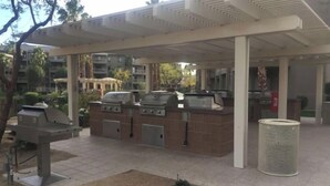 An outdoor patio area with a covered seating area, a barbecue grill, and a trash can. - Luxury 2BR Condo for Music Fests (Indio)