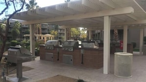 An outdoor patio area with a covered seating area, a barbecue grill, and a trash can. - Luxurious 1BR Condo for Coachella Week 2 (Indio)