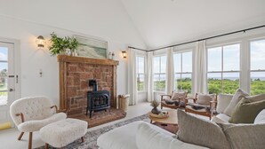 A cozy living room with a fireplace, comfortable seating, and large windows offering a view of greenery.