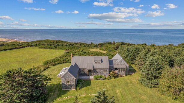 A large, two-story house with a stone exterior and a steeply pitched roof, surrounded by lush greenery and a view of the ocean.