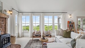 A spacious living room with a fireplace, large windows, a sofa, and a bookshelf.
