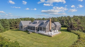 A large, modern house with a spacious patio and multiple decks, surrounded by a well-maintained lawn and dense forest.