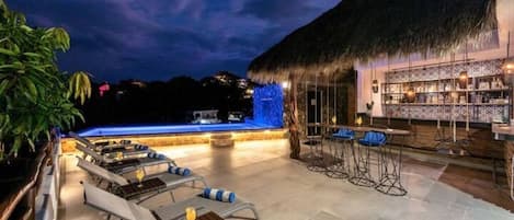 An outdoor bar area with a thatched roof, lounge chairs, and a pool with blue lighting.