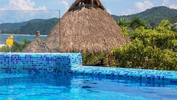 Una piscina con una estructura de techo de paja y bordeado de azulejos, rodeado de vegetación y vistas a un paisaje montañoso.