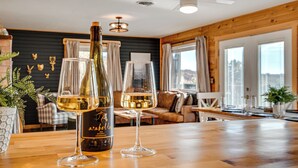 Un tavolo in legno con una bottiglia di vino Arabelle e due bicchieri, un ventilatore a soffitto in legno e un'area salotto accogliente con un divano e un tavolo da pranzo.