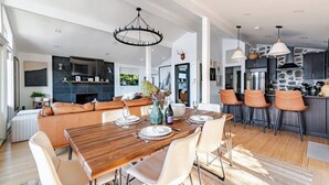 Amplia sala de estar con mesa de comedor de madera, sofás de cuero y cocina con armarios oscuros y salpicadero de piedra.