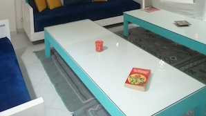 Ein Wohnzimmer mit einem weißen Sofa, einem blauen Sofa und einem weißen Sessel. Zwei Glastische und ein rotes Dekoelement stehen auf einem Tisch.