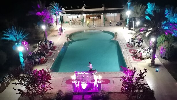 Ein nächtlicher Blick auf einen Poolbereich mit Bühne und Sitzgelegenheiten, umgeben von Palmen und beleuchtet durch farbenfrohe Lichter.