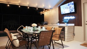 A dining area with a table set for four, surrounded by wicker chairs, a flat-screen TV mounted on the wall, and a kitchen counter with a sink and a vase of flowers. - LUXURY Villa AZUL Naples with HEATED SALT WATER POOL (Naples)