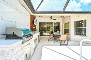 Een buitenkeuken met een grill, een eethoek met stoelen en een tafel, en een plafondventilator.