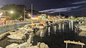 Un port de plaisance de nuit, avec des bateaux amarrés, une promenade en bord de mer et des bâtiments illuminés en arrière-plan.