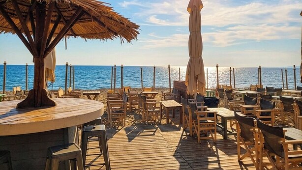Une terrasse en bois avec des tables et des chaises, un parasol en chaume et une vue sur la mer.
