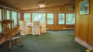 Une pièce aux murs en bois, dotée de grandes fenêtres, d’un fauteuil à bascule, de fauteuils à motifs floraux et d’une table.