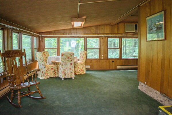 A room with wooden walls, large windows, a rocking chair, floral chairs, and a table.