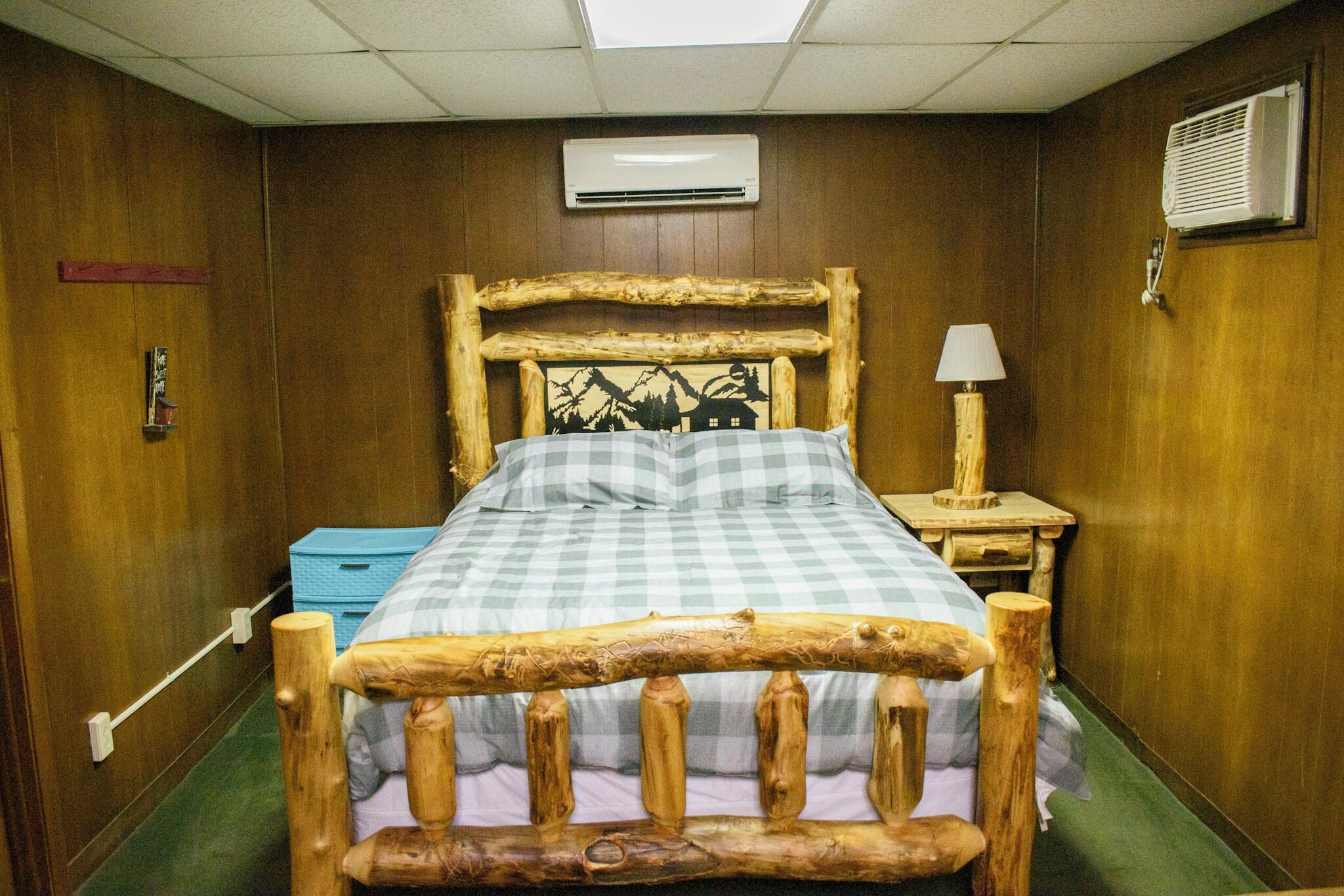 A wooden-framed bed with a checkered bedspread, a bedside lamp, and an air conditioning unit on the wall.