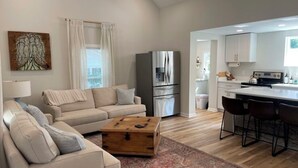 Un salón moderno con un sofá color beige, una mesa de centro de madera y una alfombra estampada. Se ve una cocina con armarios blancos y un refrigerador de acero inoxidable al fondo.