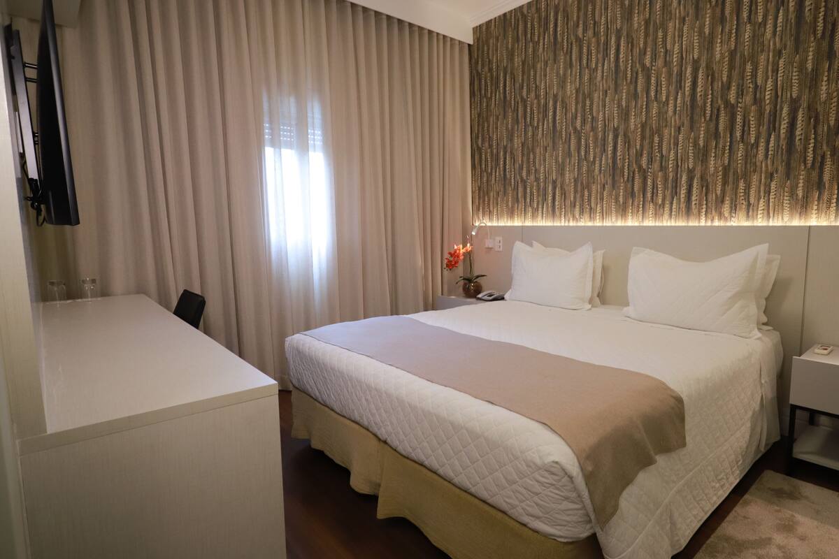 Een hotelkamer met een groot bed, een nachtkastje, een bureau en een raam met gordijnen.