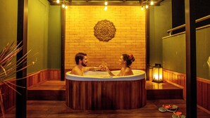 A couple in a wooden bathtub with a decorative wall behind them, a lantern, and a plant to the side. - LS Villas Tranquillità (Aguas de Sao Pedro)