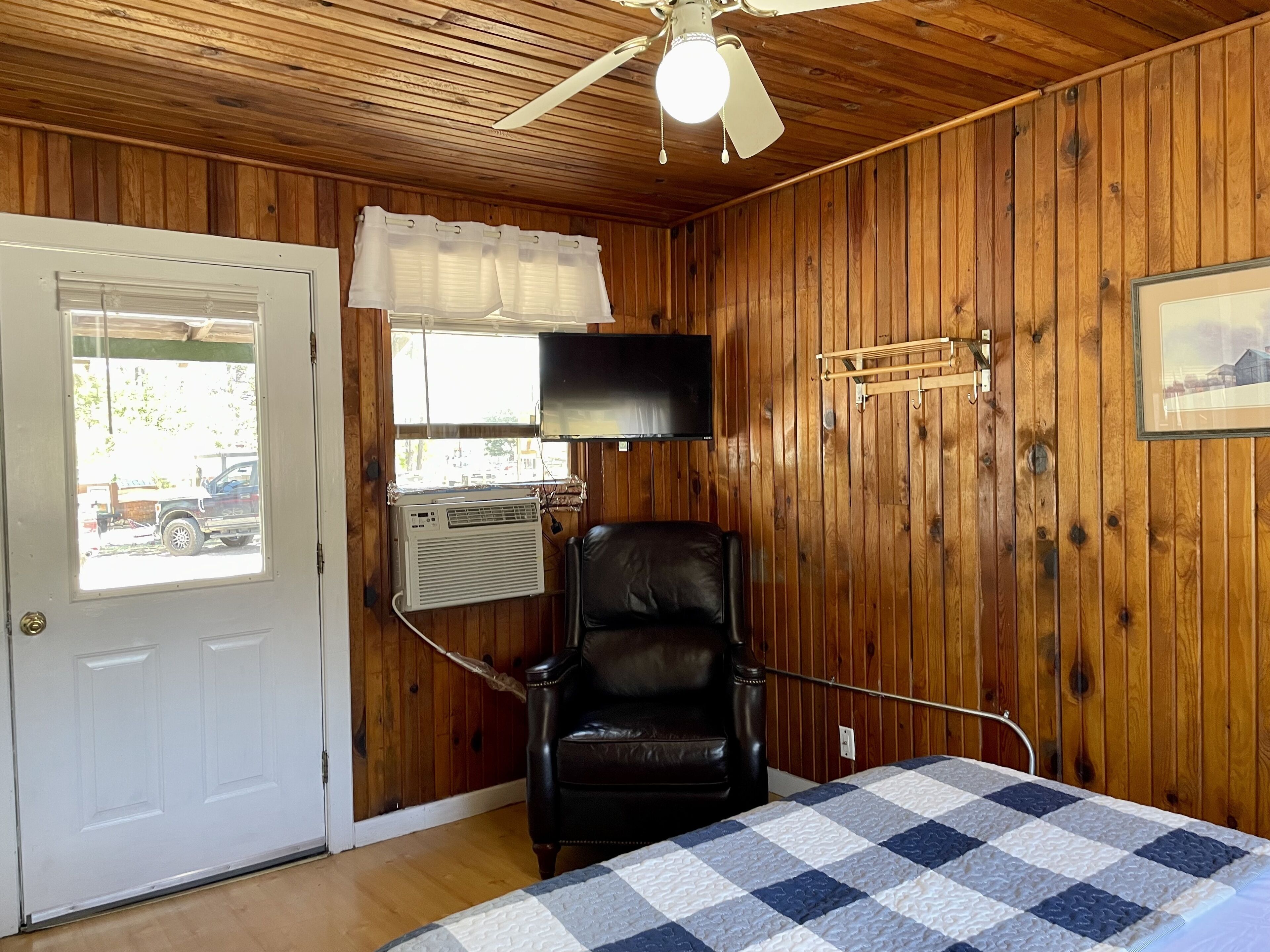 Een kamer met houten wanden, een plafondventilator, een bed met een geruit quilt, een zwarte leren fauteuil, een tv aan de muur en een raam met uitzicht op een geparkeerde vrachtwagen.